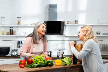 smiling woman opening wine bottle with corkscrew and looking at her asian friend