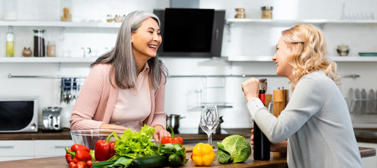 panoramic shot of smiling woman opening wine bottle with corkscrew and looking at her asian friend