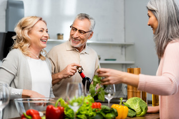 smiling woman cooking and talking with friend, man opening bottle with wine