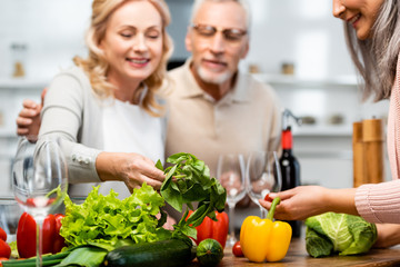 selective focus of smiling friends looking at basil in kitchen