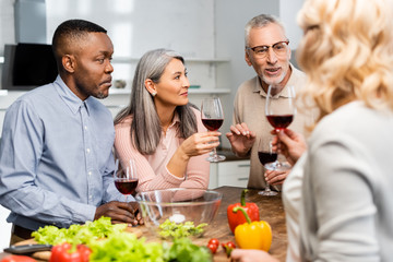 selective focus of multicultural friends talking and holding wine glasses in kitchen