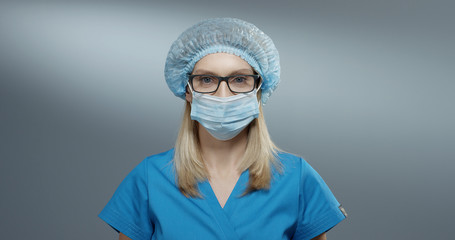 Portrait shot of the young attractive blond Caucasian female physician in the blue hat and glasses taking off her mask from the mouth and smiling happily. Close up.