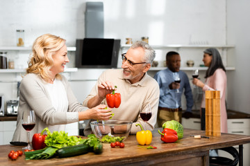 selective focus of woman adding lettuce and smiling man holding bell pepper, multicultural friends talking on background