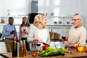 selective focus of woman cutting bell pepper and talking with man, multicultural friends talking on background