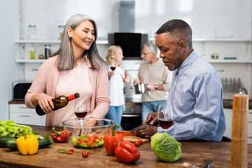 selective focus of smiling asian woman pouring wine to glass and african american man cutting bell pepper