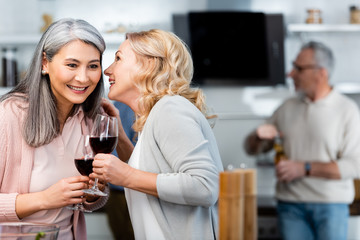 selective focus of smiling woman telling secret to asian friend with wine glass