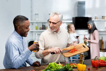 african american man holding beer and his friend adding cherry tomatoes to salad