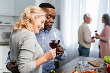 selective focus of african american man hugging and clinking with smiling friend in kitchen