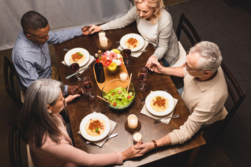 overhead view of multicultural friends holding hands and praying during dinner