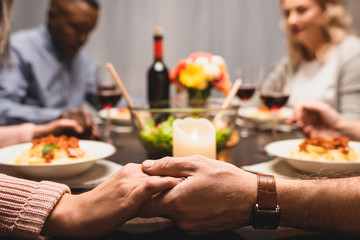 cropped view of multicultural friends holding hands and praying during dinner