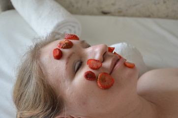 Fototapeta premium Young beautiful woman getting cosmetic fruit facial mask of fruit strawberry at beauty salon on a white towel indoors. natural cosmetic