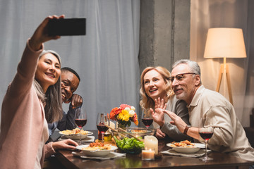 smiling multicultural friends taking selfie with smartphone during dinner
