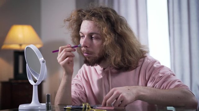 Close-up portrait of Caucasian binary gender person applying face powder and looking at mirror. Transgender doing makeup indoors. Intersex people, gender identity, self perception.