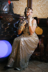 a woman in a gold dress sits on an armchair and closes with her hands in long gloves, smiling...