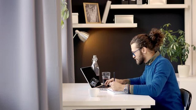 Inovative young caucasian freelancer sitting in his office, using laptop for internet search and writing in notebook results of searching.