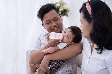 Father feeding son with milk in baby bottle, mother sitting on side