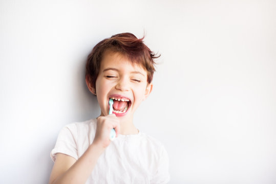 Horizontal portrait of child with closed eyes brushing teeth with blue and white toothbrush. Dental and health care from childhood. Healthy changing teeth on smiling face. Copy space for text