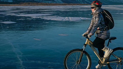 Woman is riding bicycle on the ice. Girl is dressed in a silvery down jacket, cycling backpack and helmet. Ice of the frozen Lake Baikal. Tires on bike are covered with spikes. Traveler is ride cycle.