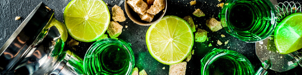 Strong alcohol. Absinthe with sugar, ice cubes and lime slices, Three shot glasses with absinthe. Dark stone background copy space
