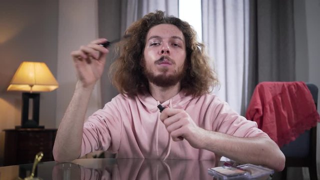 Portrait of concentrated binary gender Caucasian person applying mascara on mustaches and smiling at camera. Man with makeup on one side of face. Gender, identity, neutrality.