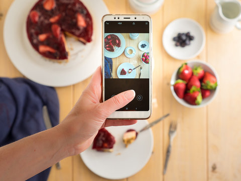 Hands hold the phone, photographing colorful, healthy food on a light wooden table. Selective focus.