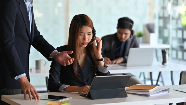 Photo of beautiful woman with headset working as call center sitting at the working desk and get coaching by her leader. - Powered by Adobe