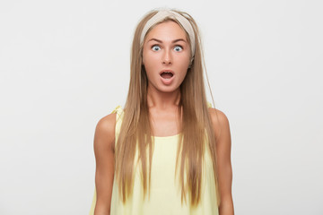 Fototapeta premium Amazed young beautiful long haired blonde female wearing beige headband while standing over white background, looking surprisedly at camera with wide eyes and mouth opened