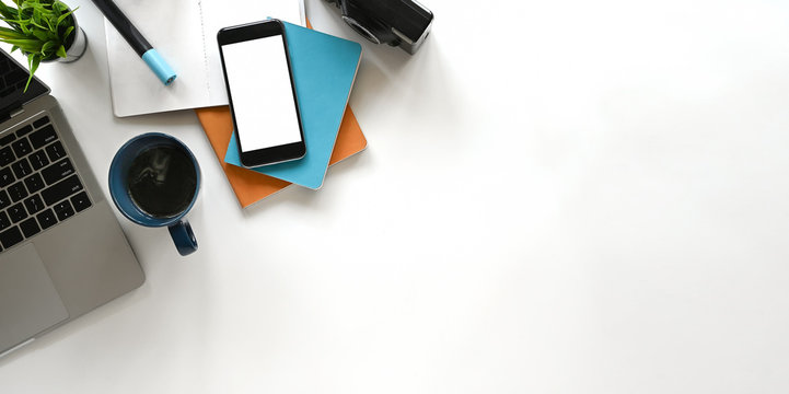 Modern office desk include laptop, camera, coffee cup, notebook, book, potted plant and pencil. Top view of white blank screen smartphone and Panorama banner background with copy space.