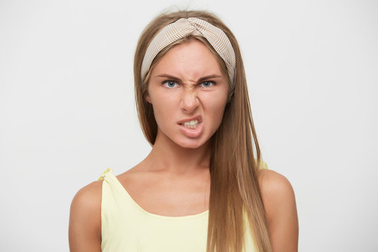 Close-up of displeased young pretty blonde lady with casual hairstyle twisting her mouth and frowning eyebrows while posing over white background, wearing beige headband and yellow top