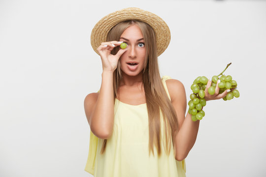 Studio photo of pretty young female with long blonde hair raising hand with grape to her eye and looking excitedly at camera, dressed in summer romantic clothes over white background