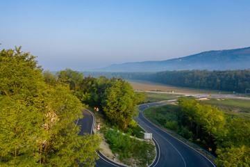A fragment of a narrow single-lane winding road with an asphalt surface. The background of the image is a mountainous area in the morning fog. The view from the top. Shooting from a drone.