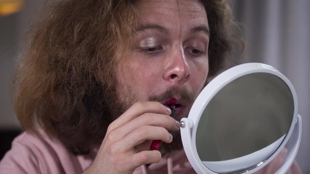 Close-up portrait of young Caucasian man with face divided into two parts applying red lipstick. Expression of intersex person self perception.