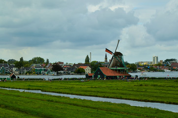 Fototapeta premium Zaanse Schans, Holland, August 2019. North-east of Amsterdam is a small community located on the quay of the Zaan river. View from above of the fields with mills, tourists are noticed. Cloudy day.