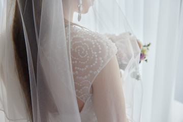 Portrait of beautiful bride in wedding dress standing with bridal bouquet near window in room, copy space. Bride's morning preparation. Wedding concept