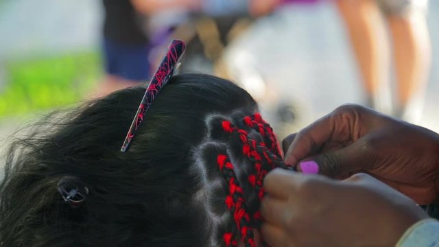 A dark-skinned girl braids pigtails with red threads to a girl.