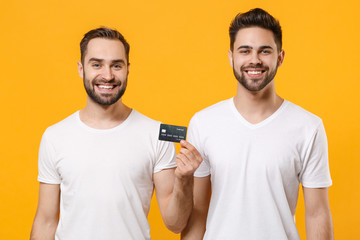 Smiling young men guys friends in white blank empty t-shirts posing isolated on yellow wall background studio portrait. People emotions lifestyle concept. Mock up copy space. Holding credit bank card.