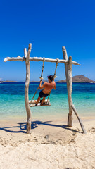 Man swinging on a wooden made swing in a very shallow water in Komodo National Park, Flores, Indonesia. There is a clear sea in front of him and some other islands in the back. Happiness and adventure