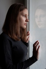 A young girl in a gray turtleneck is standing near the window her hands on the glass the reflection in the glass