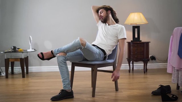 Middle shot portrait of young handsome Caucasian man sitting on chair touching hair and looking at camera. Intersex person wearing one male broggi boot and one female high heel shoe.