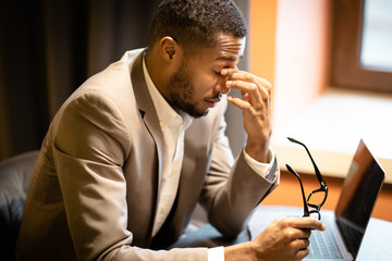Exhausted black manager massaging nose while using laptop