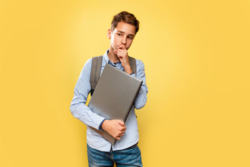 Concept of modern technologies and education. A young Caucasian teenage boy in a blue shirt and backpack, with a laptop in his hands, thoughtfully holding his chin. Yellow background. Copy space