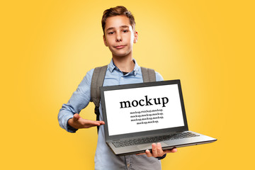 Concept of modern technologies and education. A happy Caucasian teenage boy holding an open laptop and pointing at it with his hand. Yellow background. Copy space and Mock up