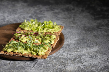 Three toast on a rye cracker with tender avocado and sprouts of micro green beetroot pea salad on a wooden Board and a gray concrete background.