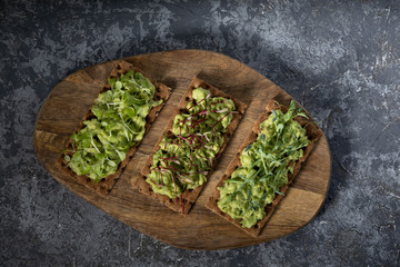 Three toast on a rye cracker with tender avocado and sprouts of micro green beetroot pea salad on a wooden Board and a gray concrete background. The view from the top