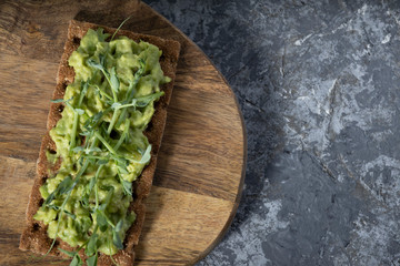 Toast on a rye cracker with tender avocado and sprouts of micro peas greens on a wooden plank and a gray concrete background. Top view