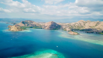 A drone shot of a paradise island with some boats anchored around in Komodo National Park, Flores, Indonesia. Brownish island turns into white sand beach and further into turquoise and navy sea.