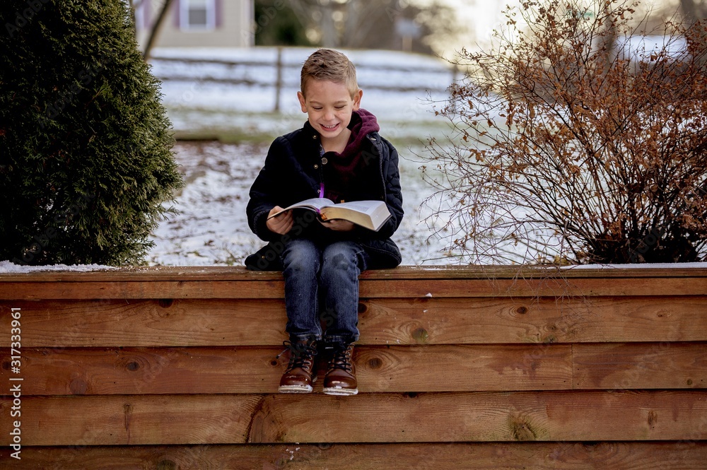 Wall mural little boy sitting on wooden planks and reading the bible in a garden covered in the snow - Wall murals