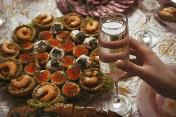 A woman holds a glass of sparkling champagne against the background of a festive table. A glass of sparkling wine in the girl's hand on the background of salads and tartlets with shrimp.