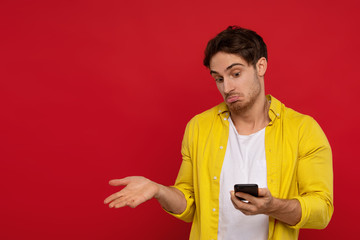 confused man in yellow shirt holding smartphone and showing shrug gesture isolated on red background