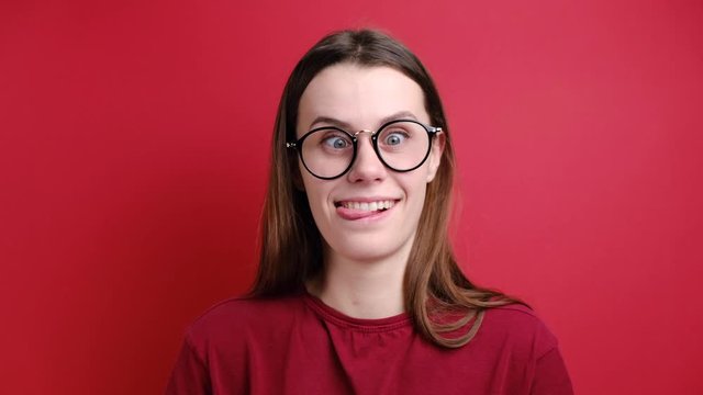 Cute young girl in glasses makes funny face, crosses eyes and sticks out tongue, plays fool, doesnt want to be responsible and looking at camera, stands over red background. Positive emotions